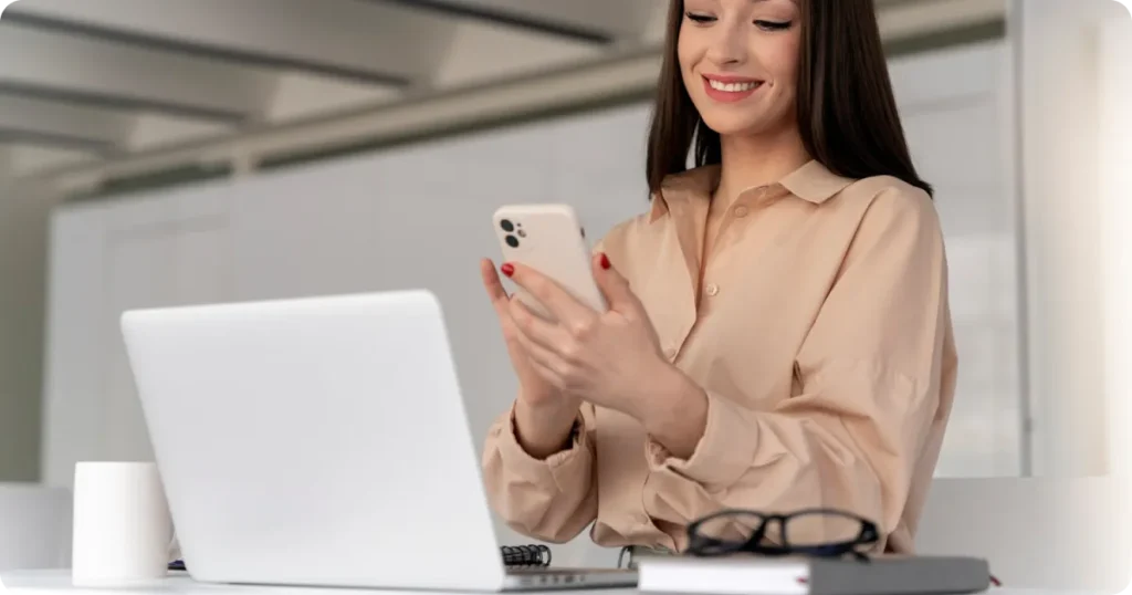 “Mulher sorrindo enquanto usa o celular ao lado de um notebook aberto, representando comunicação digital e acompanhamento de atividades de trabalho.”