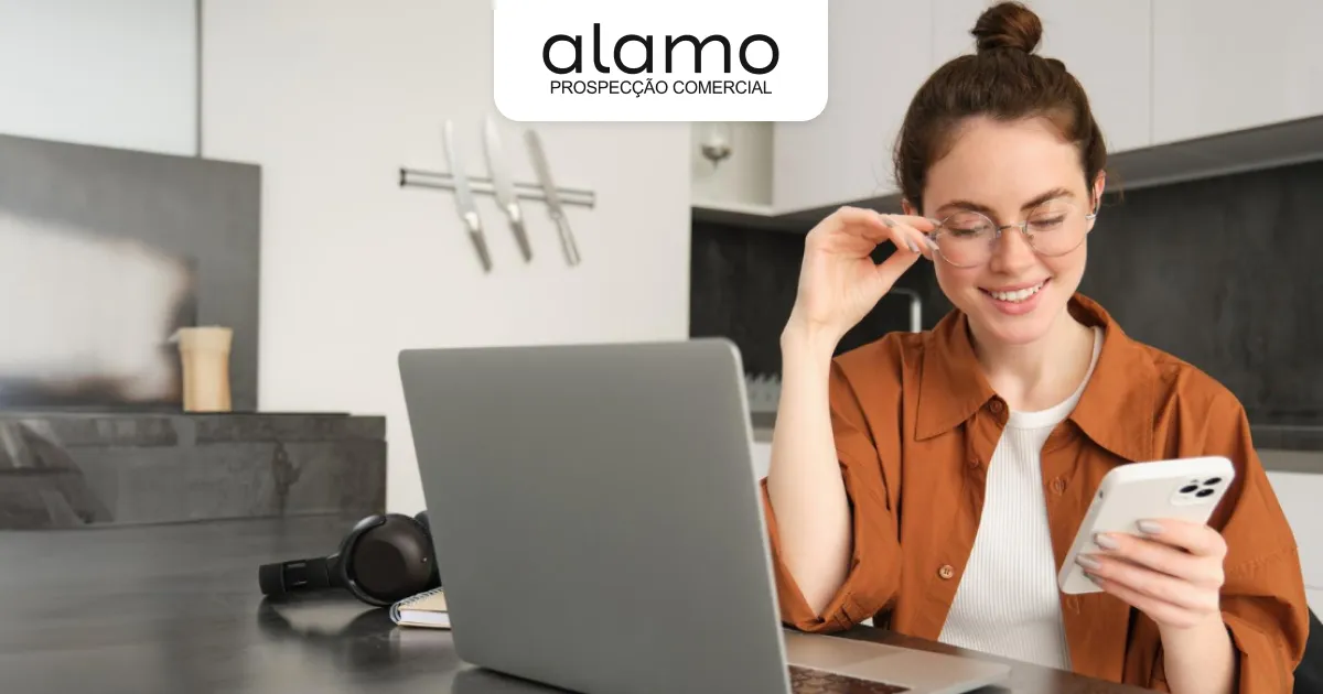 Profissional sorrindo enquanto usa o celular ao lado de um notebook, representando prospecção comercial e gestão de contatos, com logotipo da Alamo.
