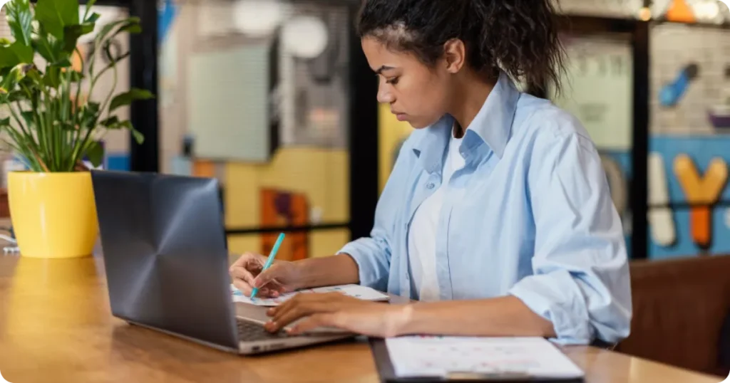 Mulher trabalhando no notebook, fazendo anotações em um caderno ao lado, em ambiente de trabalho moderno e organizado.