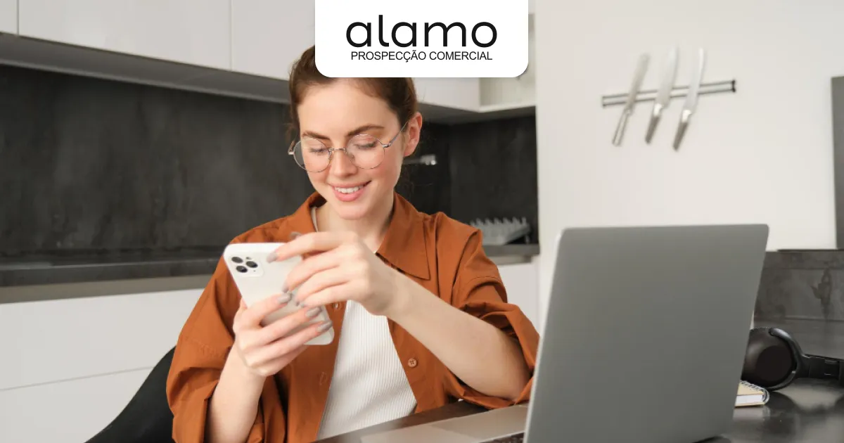 Pessoa utilizando smartphone enquanto trabalha no notebook, representando prospecção comercial e contato com clientes para geração de oportunidades de vendas.