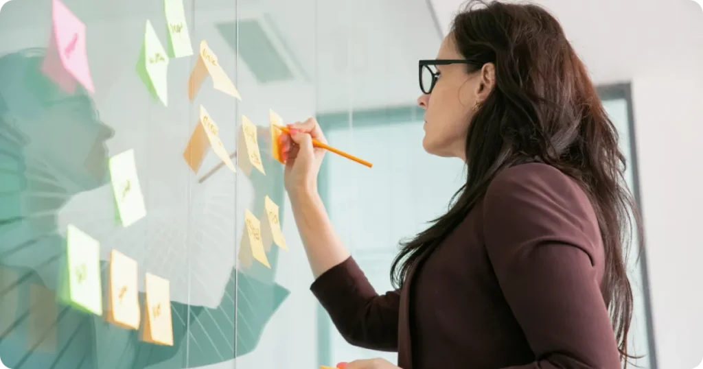 Profissional organizando ideias em um quadro com post-its, escrevendo com lápis em ambiente corporativo para planejamento estratégico.