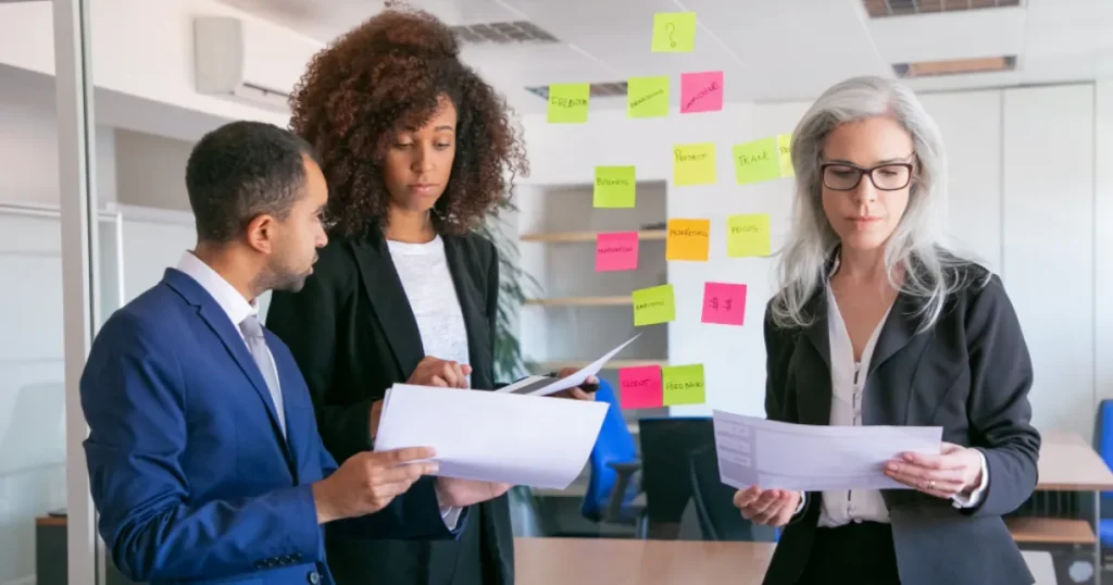 Equipe de profissionais analisando documentos em reunião, com quadro ao fundo cheio de post-its coloridos para planejamento e organização de ideias.