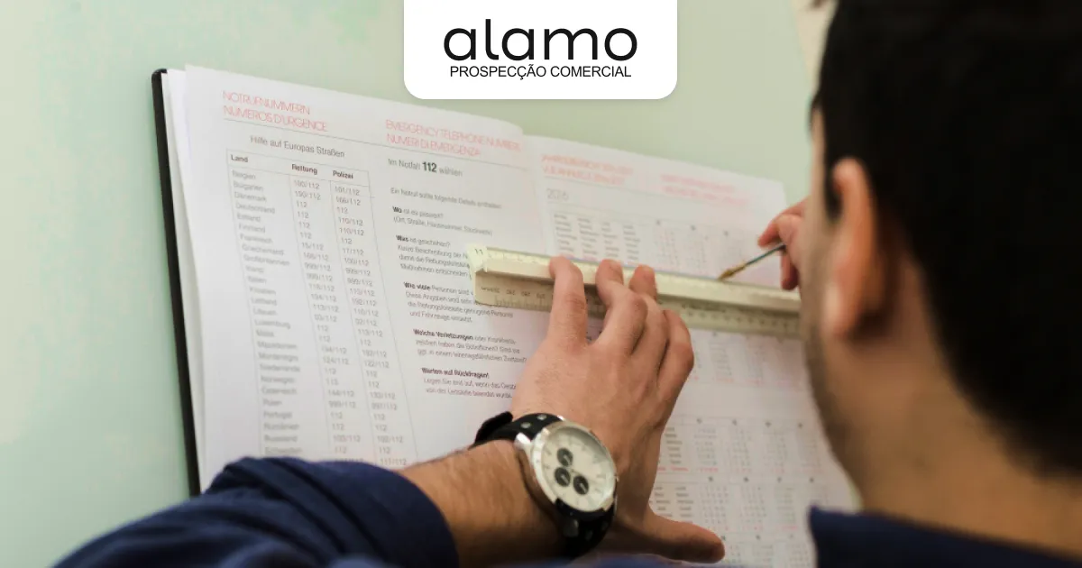 Pessoa segurando uma régua e marcando com uma caneta uma lista de um caderno aberto na mesa.