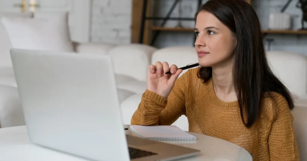 Mulher sentada em frente ao notebook, segurando um lápis enquanto pensa e faz anotações em um caderno, em ambiente doméstico.