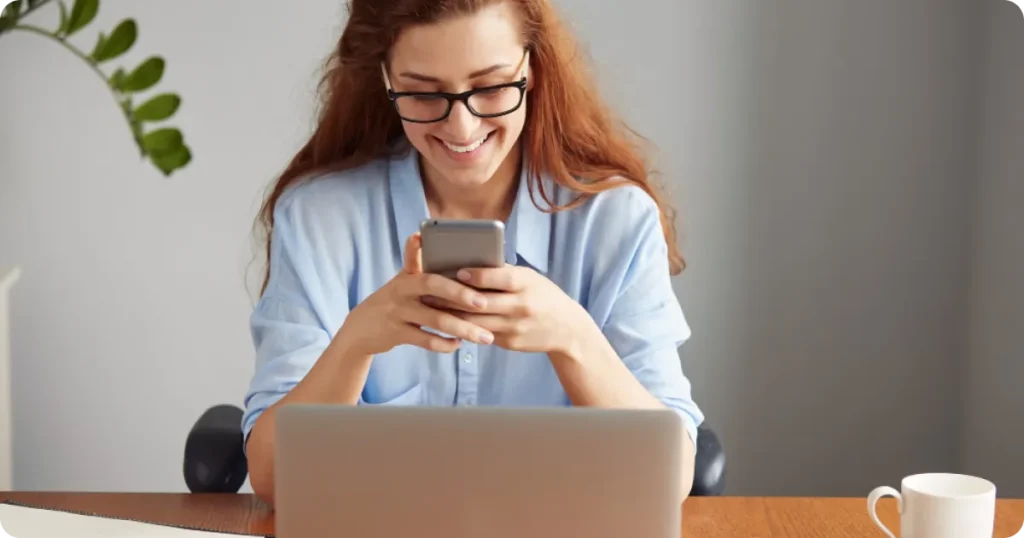Mulher sorrindo enquanto utiliza o celular em frente ao notebook, em ambiente de trabalho, demonstrando interação digital.