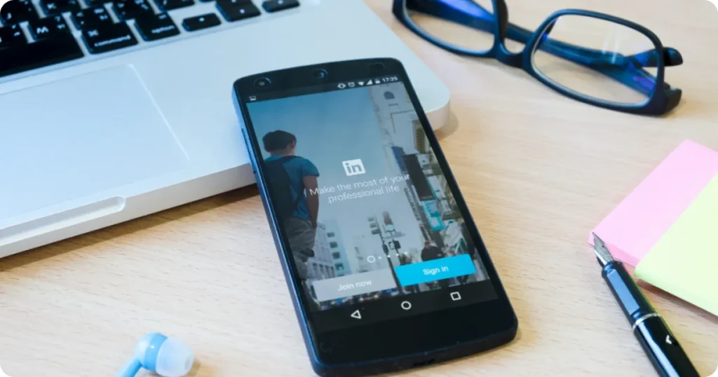 Smartphone sobre a mesa exibindo o aplicativo do LinkedIn, ao lado de notebook, óculos e materiais de escritório.